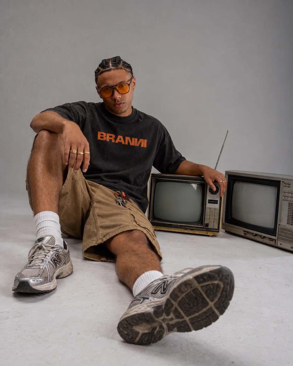 Model wearing a black and orange BRANNI T-shirt, seated beside vintage televisions